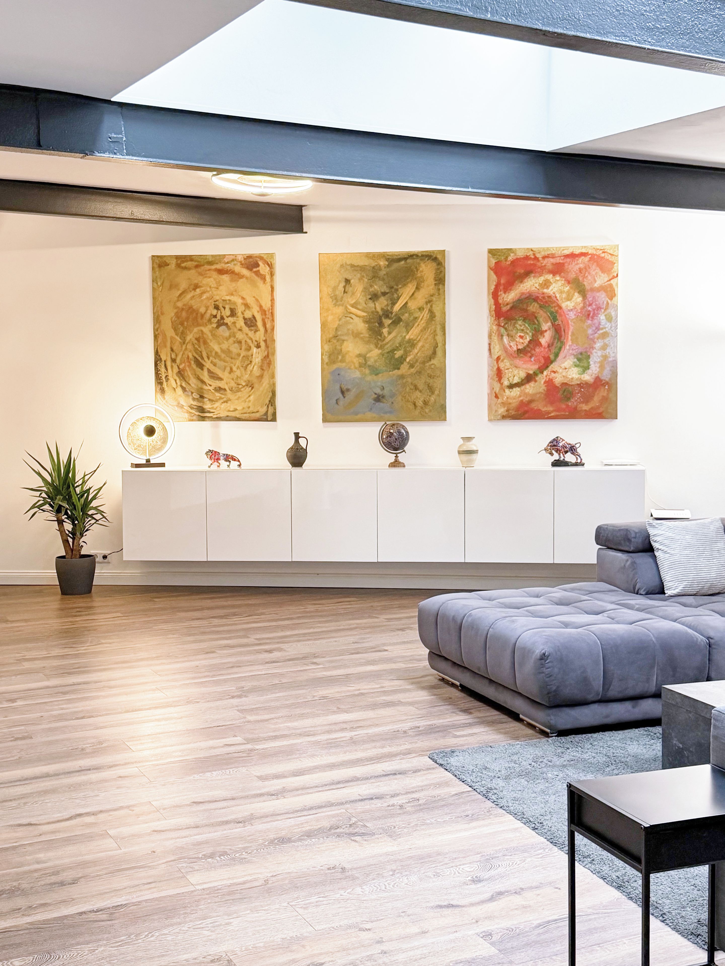 A modern living room with a sofa, a coffee table, and a side table. There are abstract paintings on the wall and a potted plant on the floor.