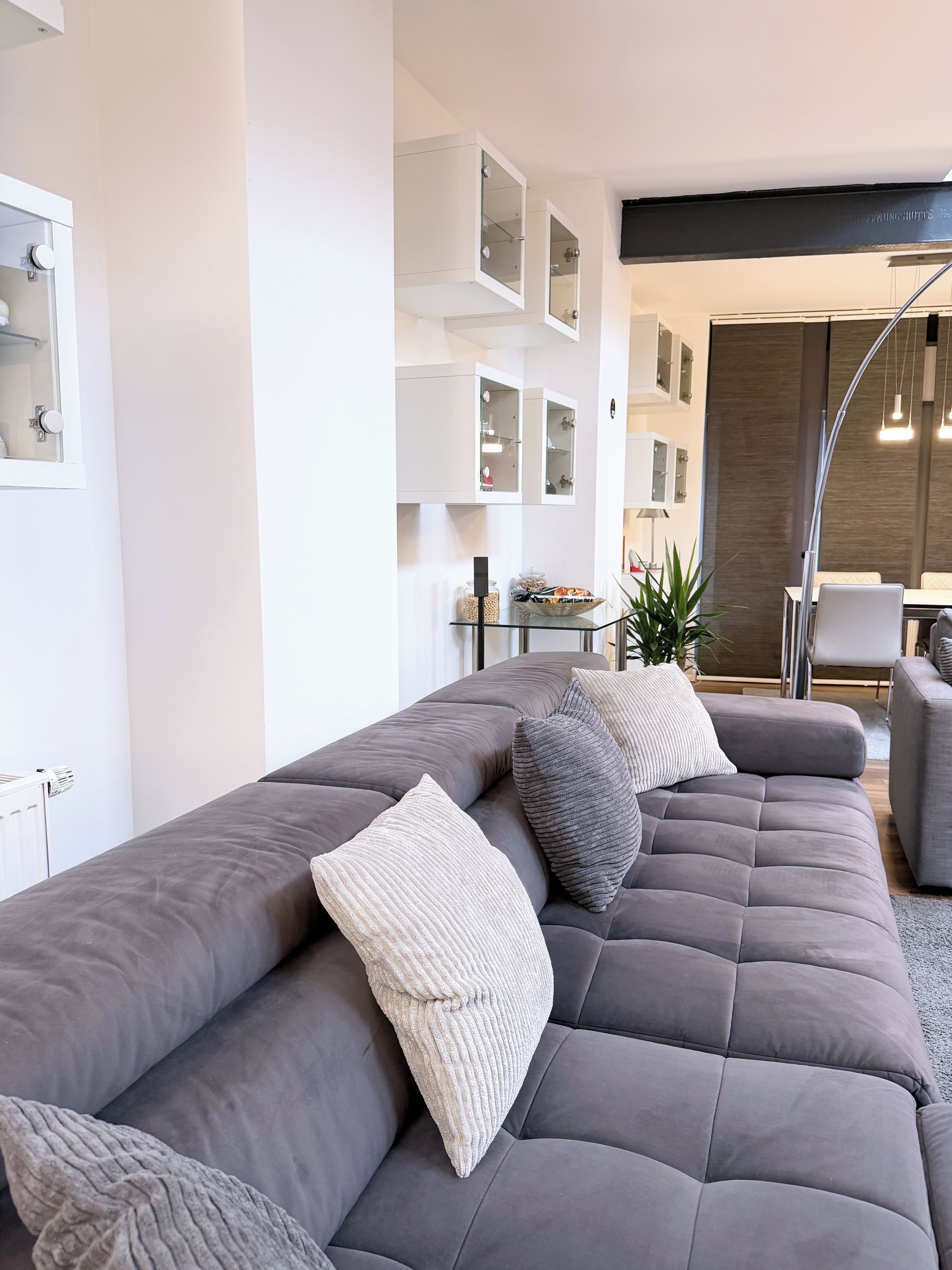 A modern living room with a grey sectional sofa, a glass coffee table, and a dining area with a white table and chairs.