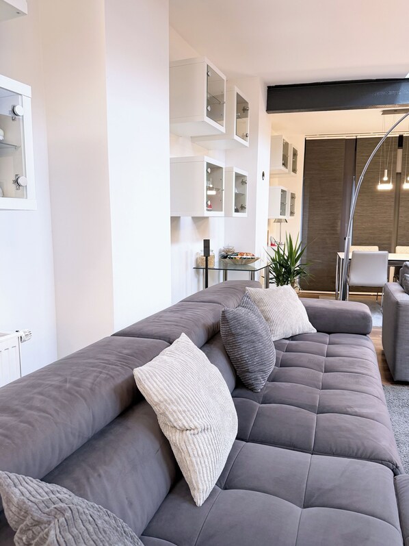 A modern living room with a grey sectional sofa, a glass coffee table, and a dining area with a white table and chairs.
