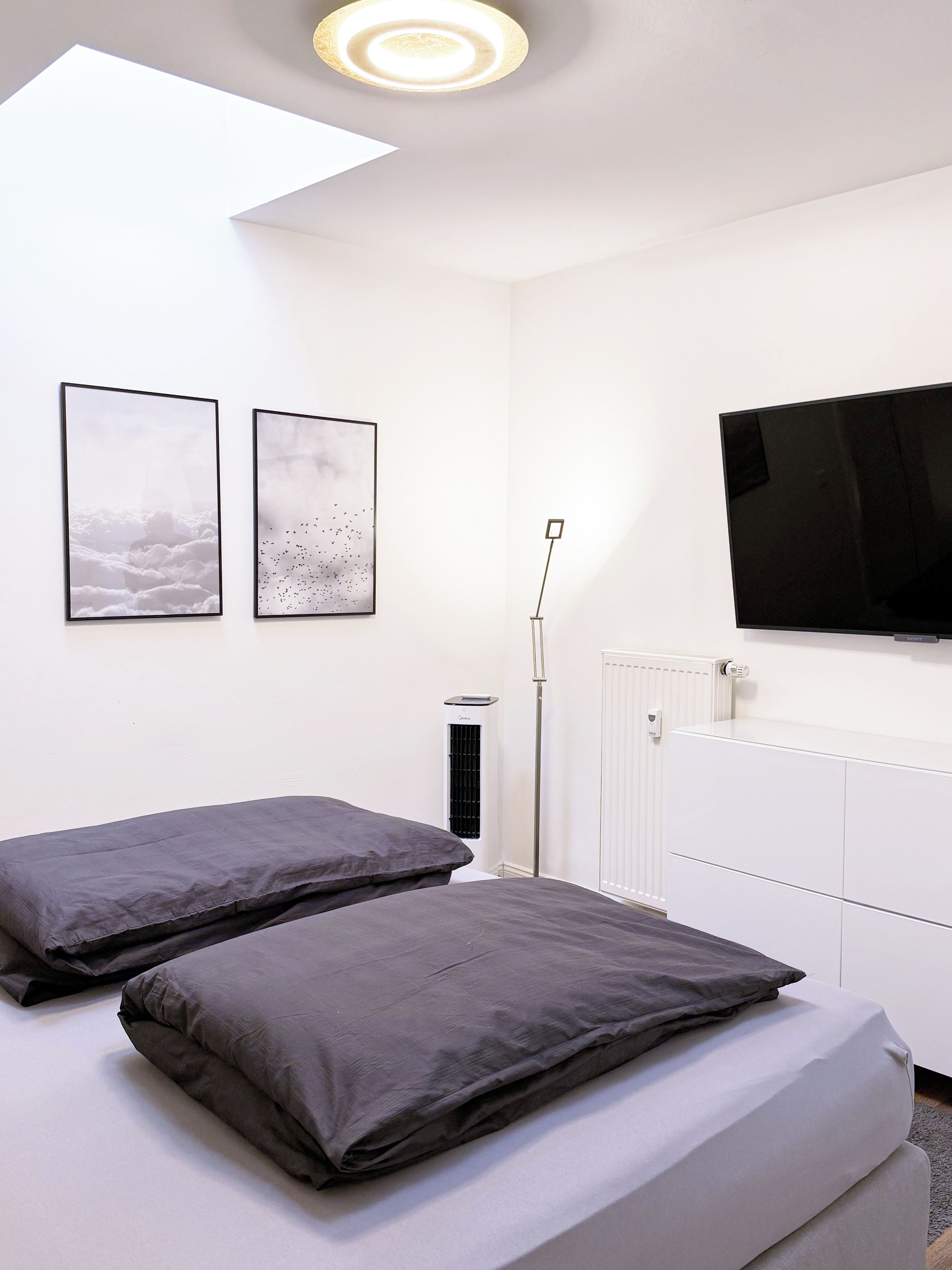 A bedroom with two beds, a ceiling light, a radiator, and a TV mounted on the wall.