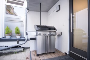Una parrilla de acero inoxidable con cuatro quemadores, ubicada sobre una terraza de madera junto a un edificio blanco con puerta de cristal.