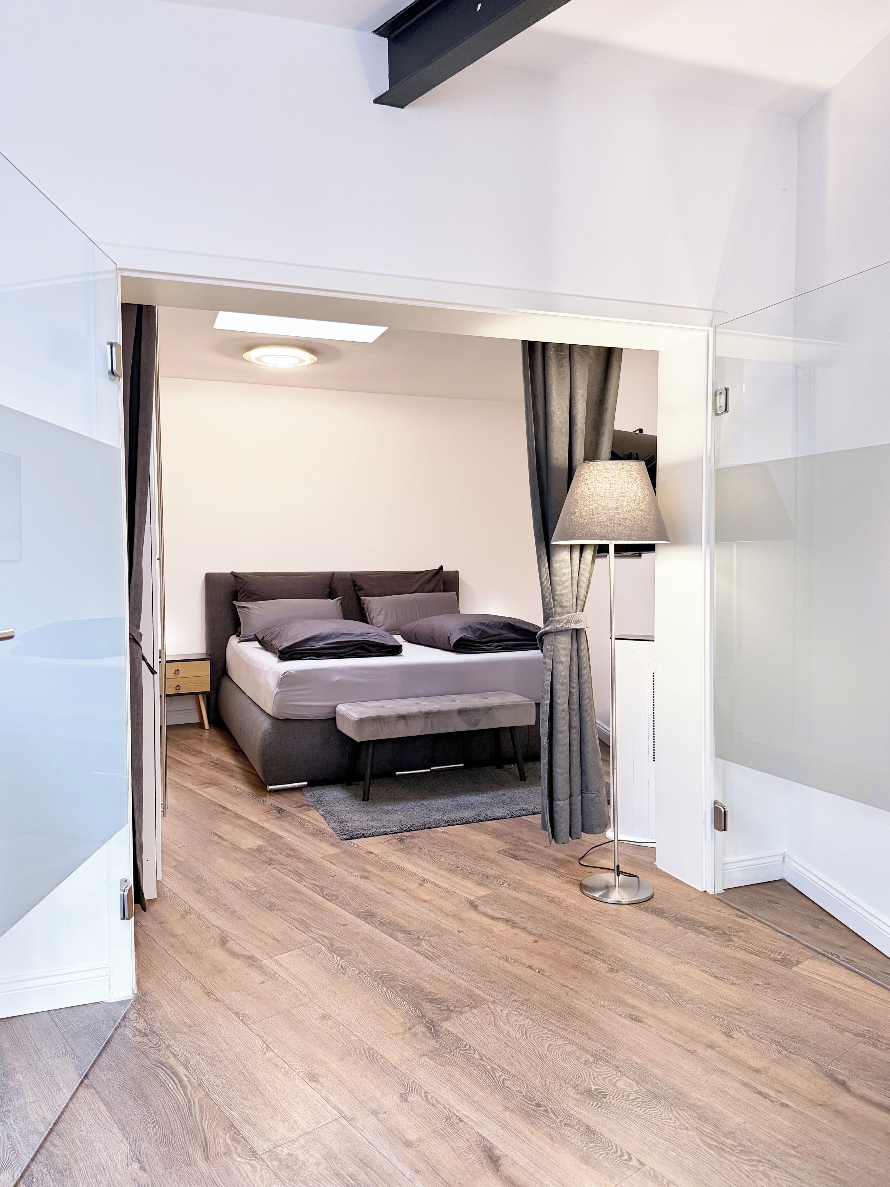 A modern bedroom with a bed, bedside table, lamp, and a view into another room.