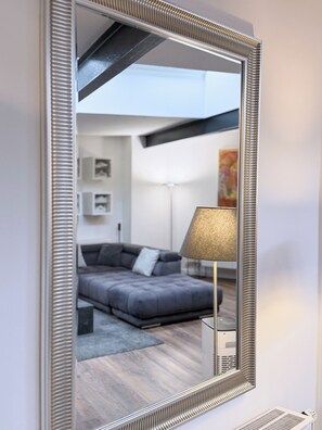 Un salon moderne avec un grand miroir, un canapé gris, une lampe sur pied et un plafond avec des poutres apparentes.