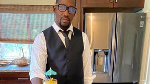 Una persona en una cocina sosteniendo dos bebidas con sombrillas, una bandeja con comida y utensilios, y un refrigerador al fondo.