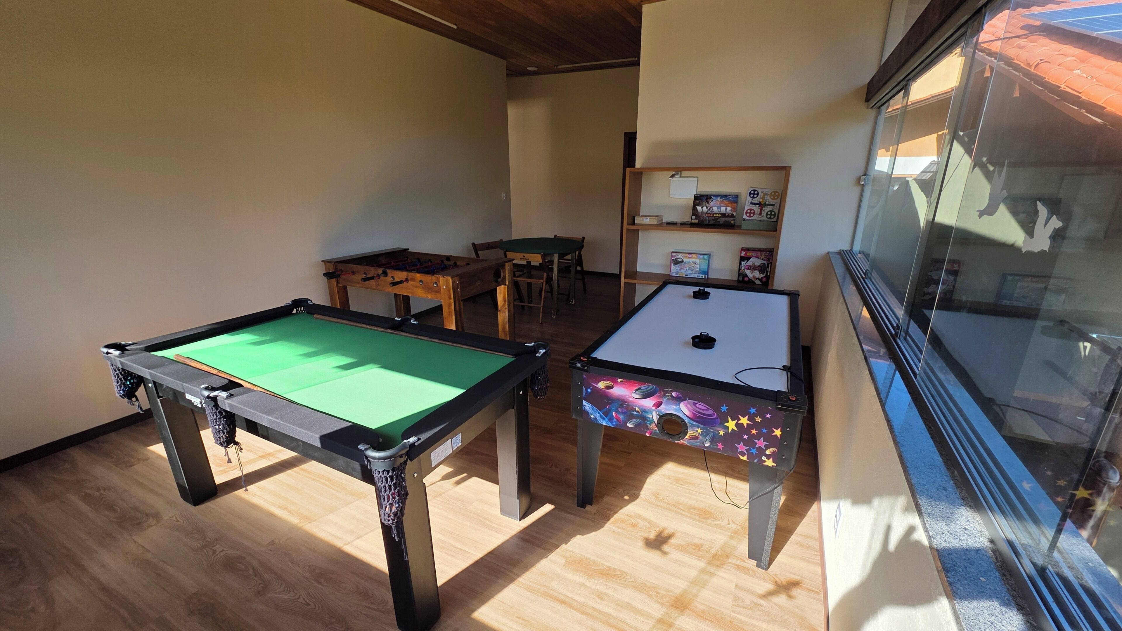 Una mesa de billar con superficie de fieltro verde, una mesa de fútbol de mesa con diseño de temática espacial, y una mesa de madera con sillas en una habitación con suelo de madera y una ventana.