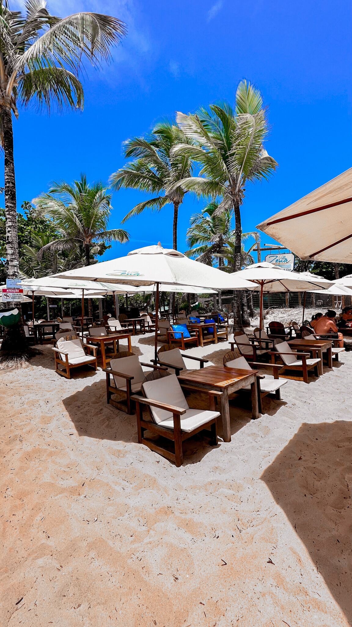 A beachside café with wooden tables and chairs, umbrellas, and palm trees.