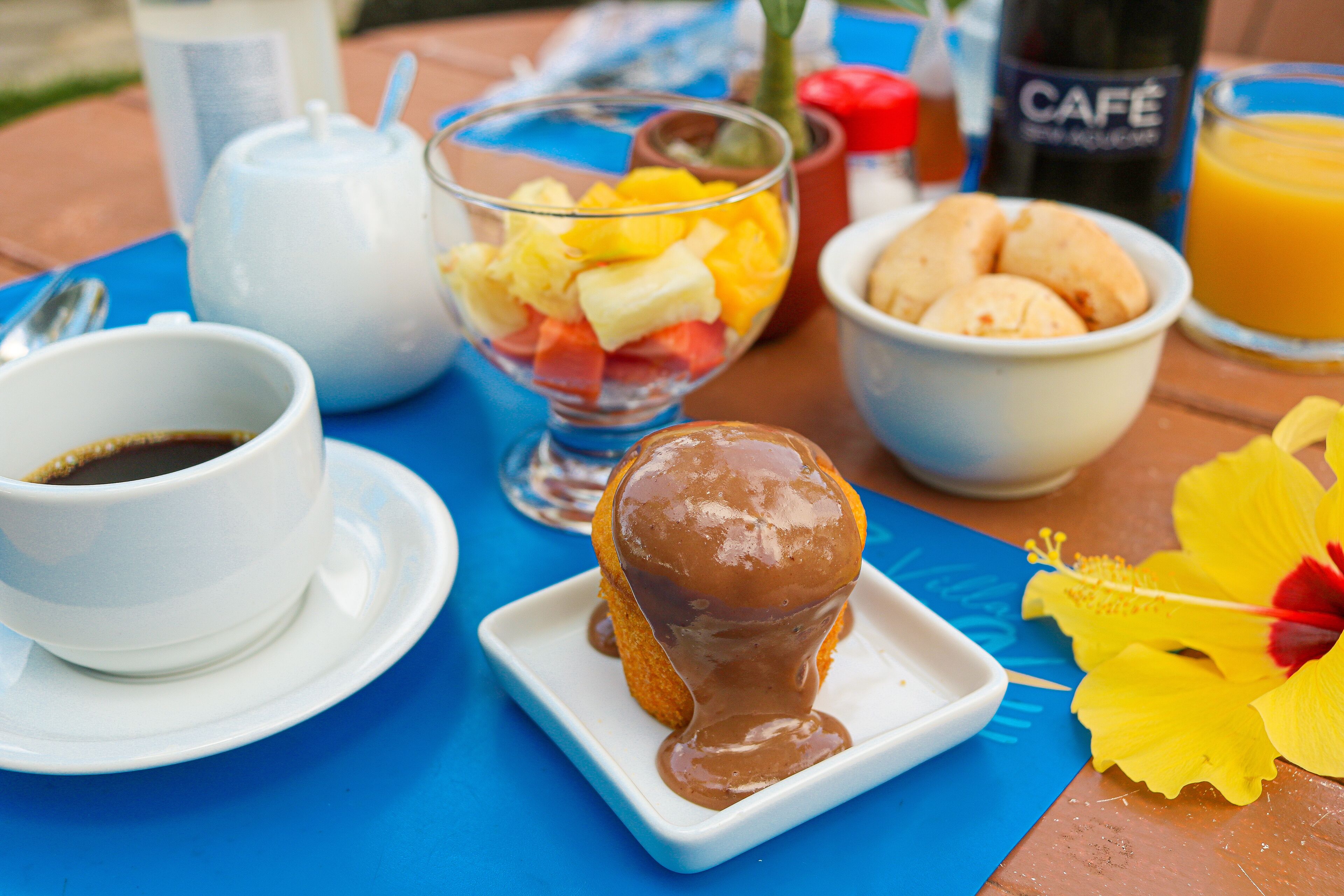 A breakfast table with a cup of coffee, a glass of orange juice, a bowl of fruit, a pastry with chocolate sauce, and a flower.