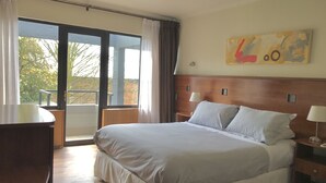 A hotel room with a large bed, a desk, a chair, a balcony with a view of trees, and a painting on the wall. - Hotel Turismo Temuco (Temuco)