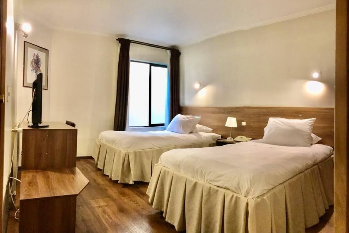A hotel room with two beds, a wooden desk, a telephone, and a window with curtains.