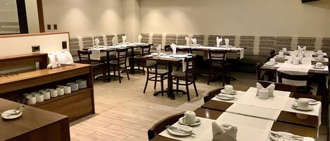 A restaurant with multiple dining tables set with white tablecloths and napkins, wooden chairs, and a buffet area with stacked plates.
