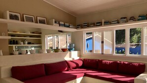 Une pièce comprenant un canapé d’angle rouge, une console blanche ornée de livres et d’objets décoratifs, et de grandes fenêtres donnant sur l’extérieur.