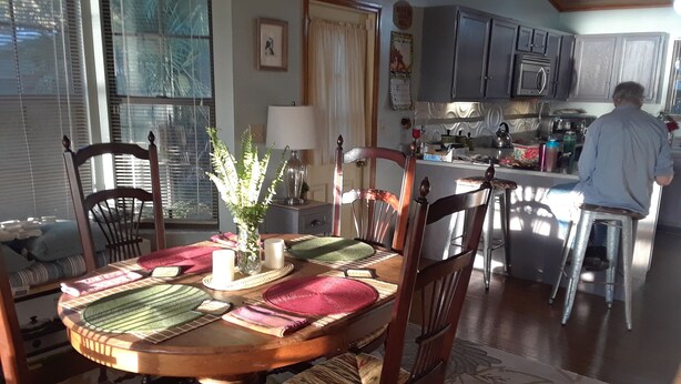 A dining area with a round table, chairs, a vase with greenery, and a kitchen in the background. - Peaceful Lakefront Getaway, Stunning Sunsets & Waterfront Relaxation (Keystone Heights)
