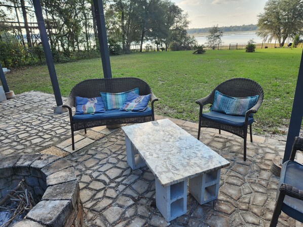 An outdoor seating area with two wicker sofas and a stone table, surrounded by a stone patio and a grassy lawn with trees and a lake view. - Peaceful Lakefront Getaway, Stunning Sunsets & Waterfront Relaxation (Keystone Heights)