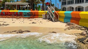 A beach with a colorful fence, a bar named 'ANDBAR', and a building with balconies.