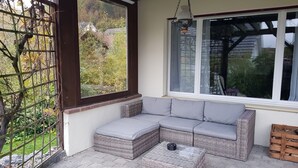 A covered patio with a wicker sectional sofa, a small table, and a hanging lantern. - Holiday home Luma (with terrace and garden) (Egerkingen)