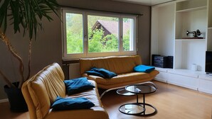 A living room with two yellow leather sofas, a glass coffee table, and a large window with a view of a house and trees. - Holiday home Luma (with terrace and garden) (Egerkingen)