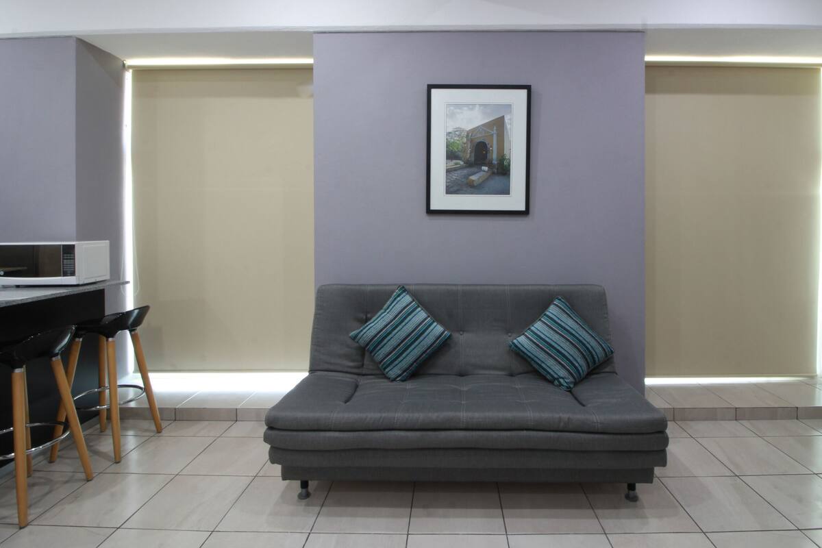 A modern office space with a grey sofa, two striped pillows, a framed picture on the wall, a white microwave on a counter, and a chair with wooden legs.
