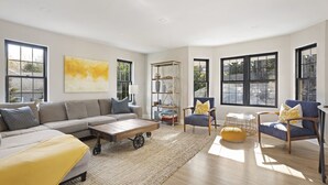 Amplia sala de estar con un sofá grande, una mesa de centro y dos sillones. Varias ventanas ofrecen luz natural y vistas al exterior.