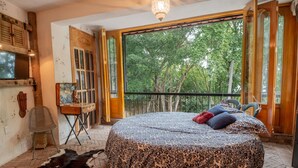 A room with a large bed, a wooden desk, and a view of trees through a balcony door.