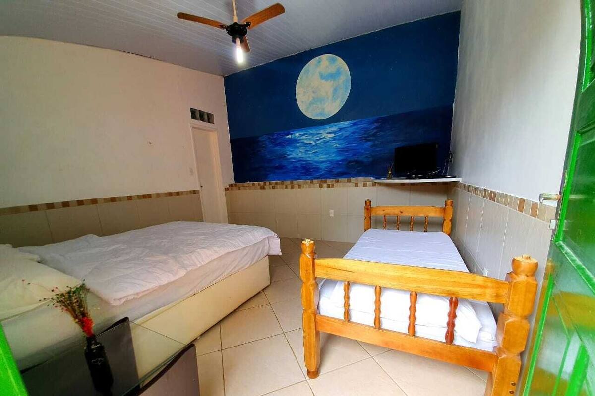 Une chambre avec deux lits, l’un en bois et l’autre en métal, un ventilateur de plafond, une fresque murale représentant la lune et l’océan, et une porte verte.