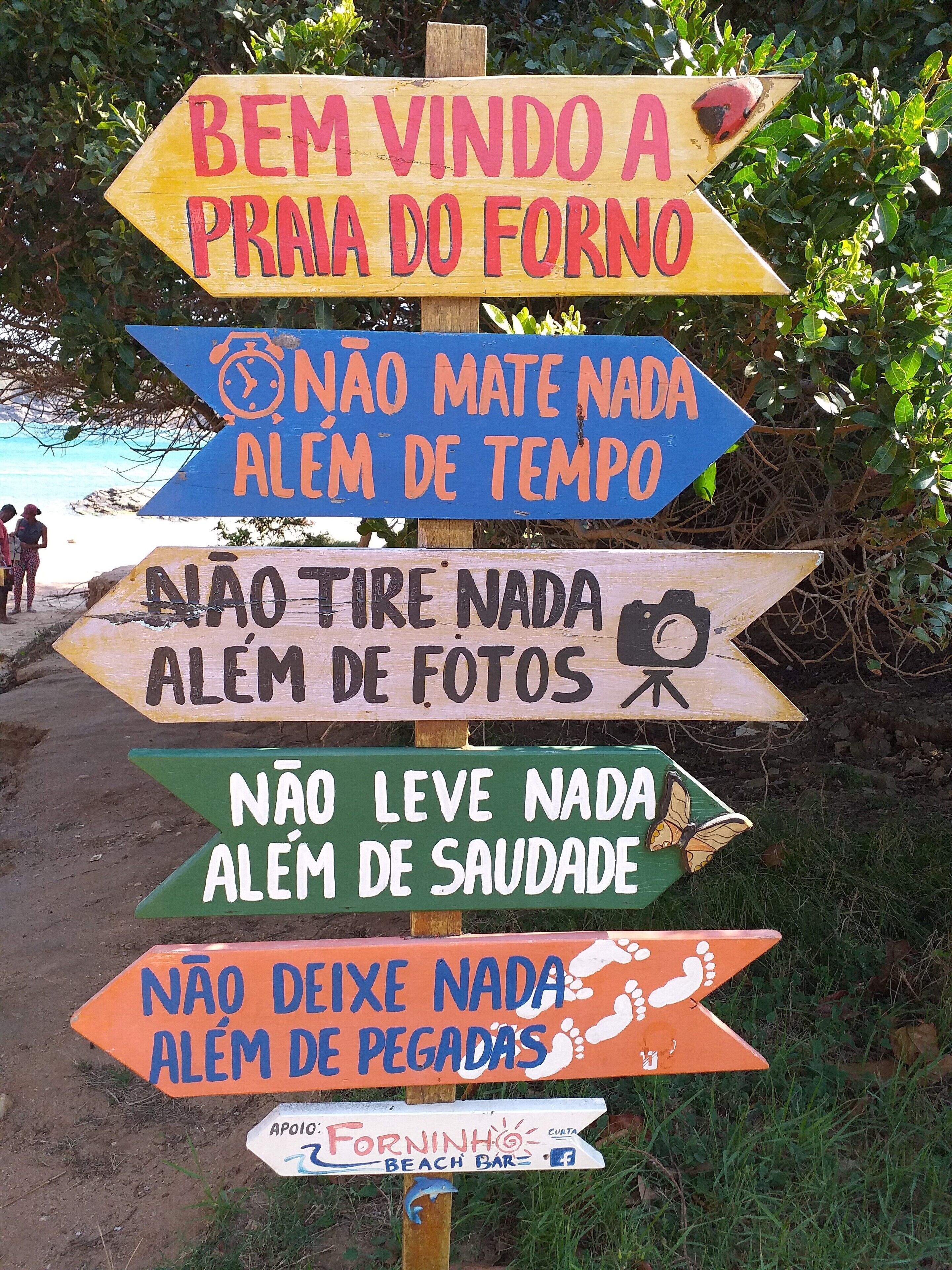 A wooden sign with multiple directional arrows and messages in Portuguese, including one pointing to a beach bar.