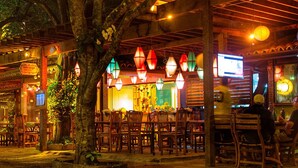 An outdoor dining area with wooden tables and chairs, surrounded by trees and illuminated by hanging lanterns. - Hostel Minha Lua (Búzios)
