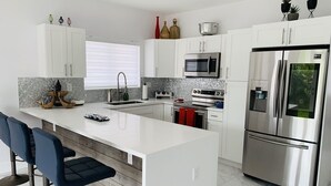 A modern kitchen with white cabinetry, a stainless steel refrigerator, a microwave, and an oven. There are two bar stools with blue upholstery, a large island with a sink, and decorative items on the cabinets.
