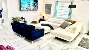 A living room with a white sofa, blue armchairs, a TV on the wall, a plant, and a window with curtains.