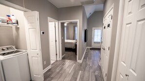 Un área de lavandería con lavadora, un estante con productos de limpieza y un pasillo que conduce a un dormitorio con cama y cortinas.