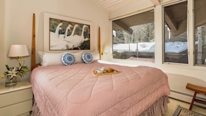 A bedroom with a large bed, a painting of swans, a window with a view of trees, and a bedside table with a lamp and a vase of flowers.