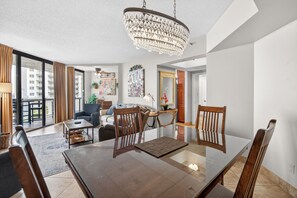 A modern living room with a glass dining table, wooden chairs, a chandelier, and a view of a cityscape through large windows.