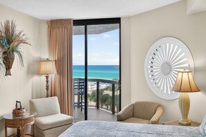 Habitación de hotel con una cama, dos sillas, una mesita, una lámpara, una planta en maceta y un ventanal con vistas al océano.