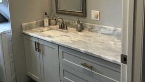 A bathroom with a marble countertop, a large mirror, a sink with a faucet, and a soap dispenser. There are cabinets with drawers and a door with a handle.