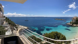 Vista costanera des d'un balcó amb mar blau clar, una platja sorrenca i edificis al fons.