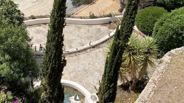 Una platja amb aigua turquesa cristal·lina, un tram de sorra i poques persones. Hi ha cypresses alts i un edifici blanc amb una piscina.