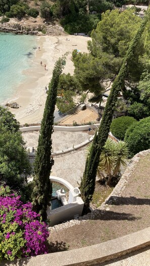 Uma praia com águas turquesa cristalinas, areal arenoso e algumas pessoas. Há ciprestes altos e um edifício branco com piscina.