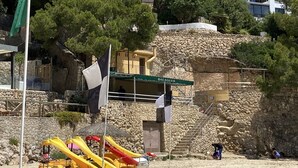 Una platja amb pedaleres grogues i vermelles, una paret de pedra i edificis al fons.