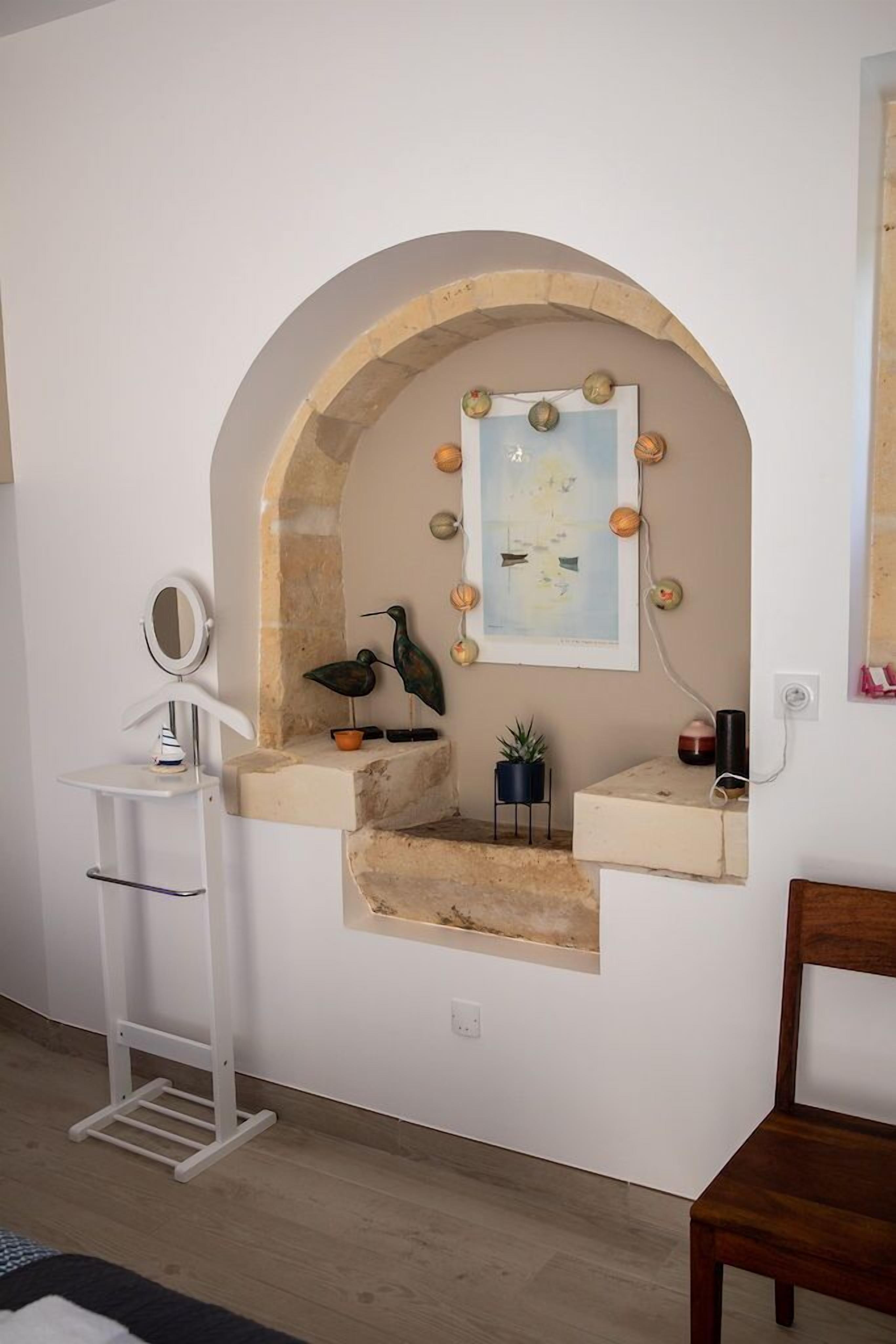 A bathroom with a stone arch, a mirror, a sink, a small shelf with decorative items, and a framed picture on the wall.