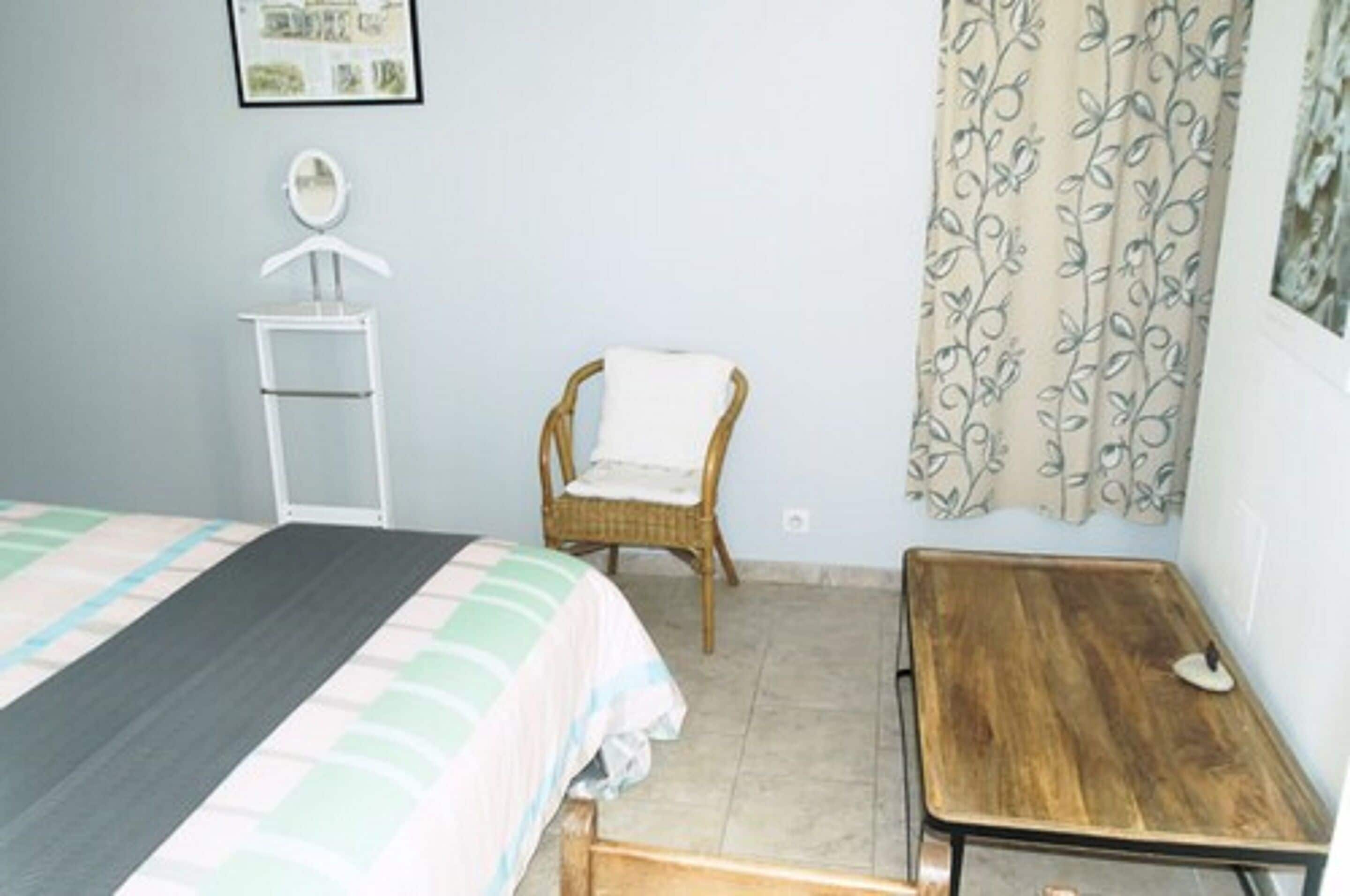 Un dormitorio con una cama, una silla, una mesita de madera, un espejo y una cortina.