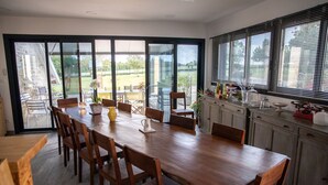 A dining area with a large wooden table and chairs, a kitchen with cabinets and a window with blinds, and a view of a garden through glass doors. - Best Saintonge (Port-d'Envaux)