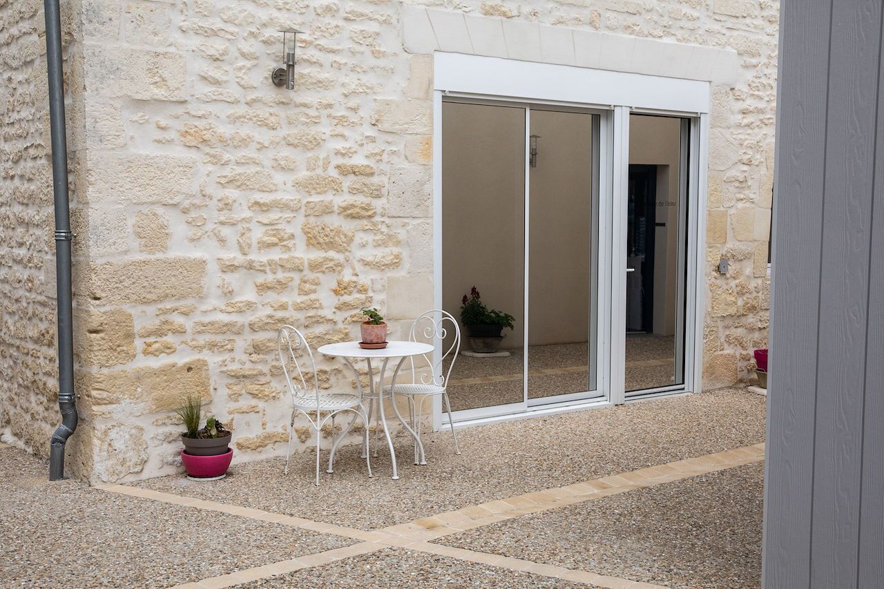 Un bâtiment en pierre doté d’un patio comprenant une table et des chaises, des plantes en pot et une porte-fenêtre coulissante donnant sur un espace intérieur.
