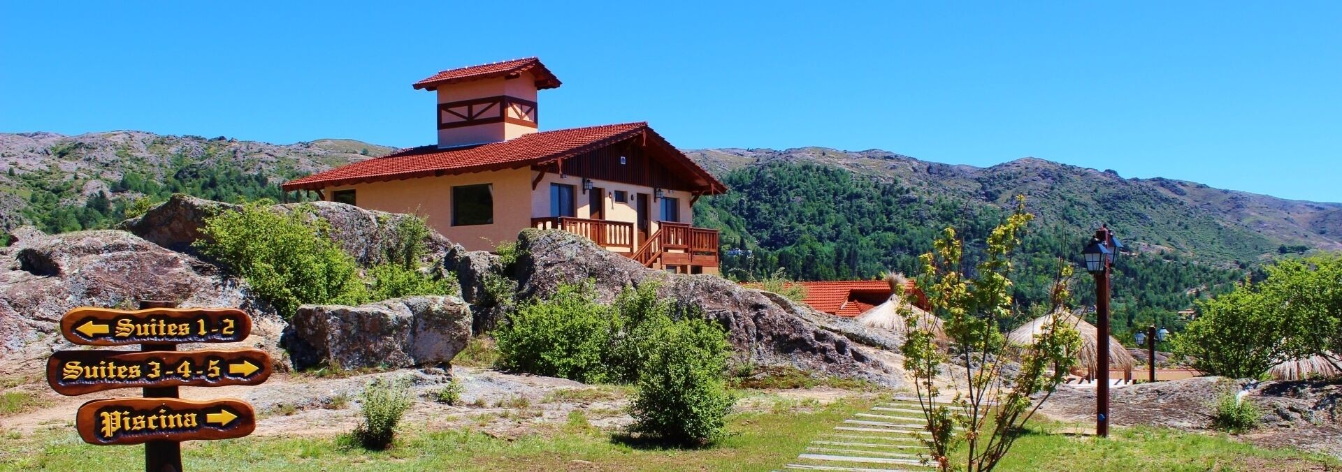 Una cabaña de montaña con techo de tejas rojas, rodeada de terreno rocoso y vegetación. La señalización indica varias suites y una piscina cercana.