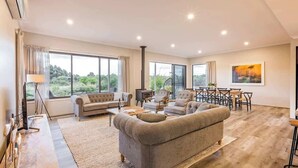 A spacious living room with a large sofa, a dining area, and large windows offering a view of greenery. - Piper—Family Retreat in the Heart of Wine Country (Wilyabrup)