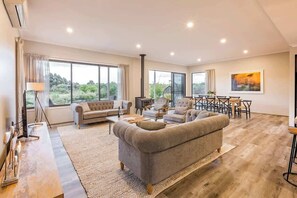 A spacious living room with a large sofa, a dining area, and large windows offering a view of greenery.