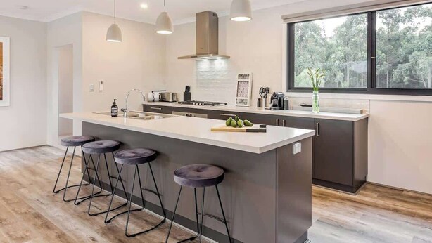 Una cocina moderna con una isla central, taburetes de bar, iluminación colgante y un amplio ventanal con vista a  árboles.