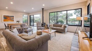 Amplia sala de estar con sofá, sillones, una mesa de centro y televisor.  Se observa una mesa de comedor con sillas y un amplio ventanal que ofrece vistas al exterior.