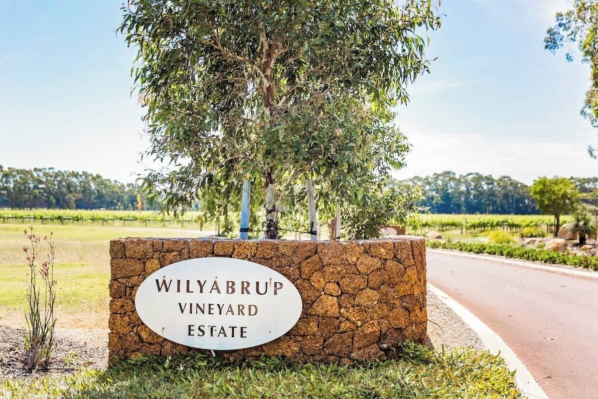 A sign for Wilyabrup Vineyard Estate with a stone wall and a tree in the foreground.