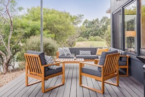 Une terrasse avec un canapé et des fauteuils, une table avec des verres et une bouteille, et une vue sur des arbres et un bâtiment.