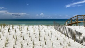 A beach with a wooden walkway leading to a wooden structure, surrounded by white sand and sparse vegetation.