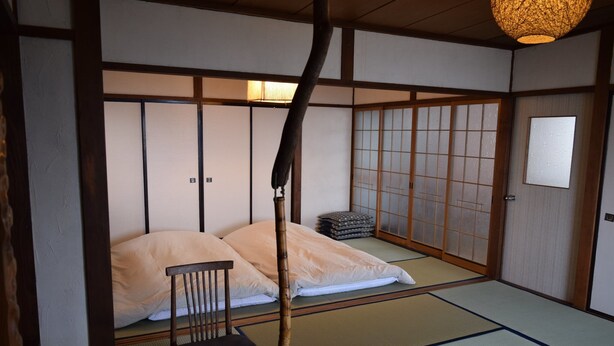 Ein traditioneller japanischer Raum mit Tatami-Bodenbelag, einem niedrigen Holzbett mit Kissen, einem Holzstuhl und verschiebbaren Shoji-Türen.